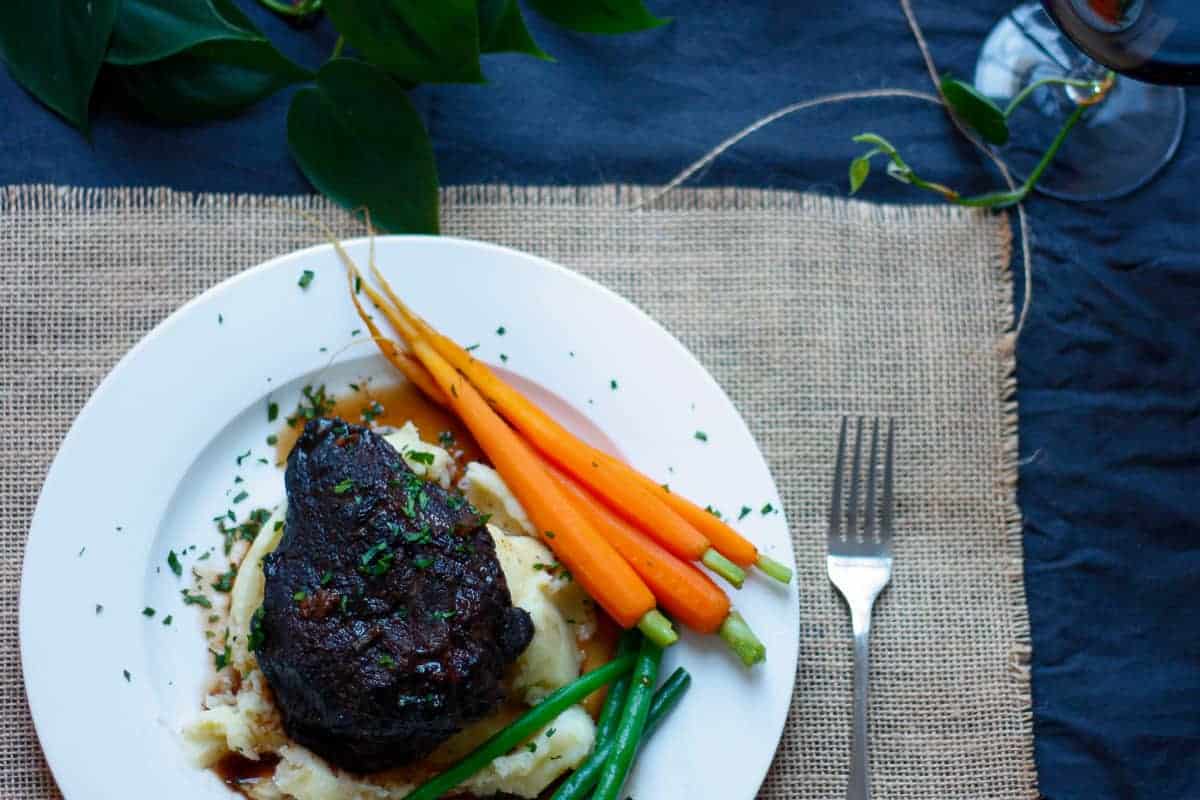 Overhead view of a whole beef cheek atop a bed of mashed potato. with baby carrots and steamed green beans on a white plate.