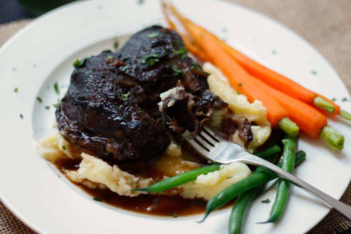 Braised beef cheek atop a bed of mashed potato with baby carrots and steamed green beans on a white plate.