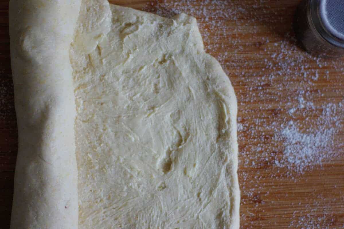 Rectangle of dough spread with butter, partially rolled up on a floured wooden work surface.