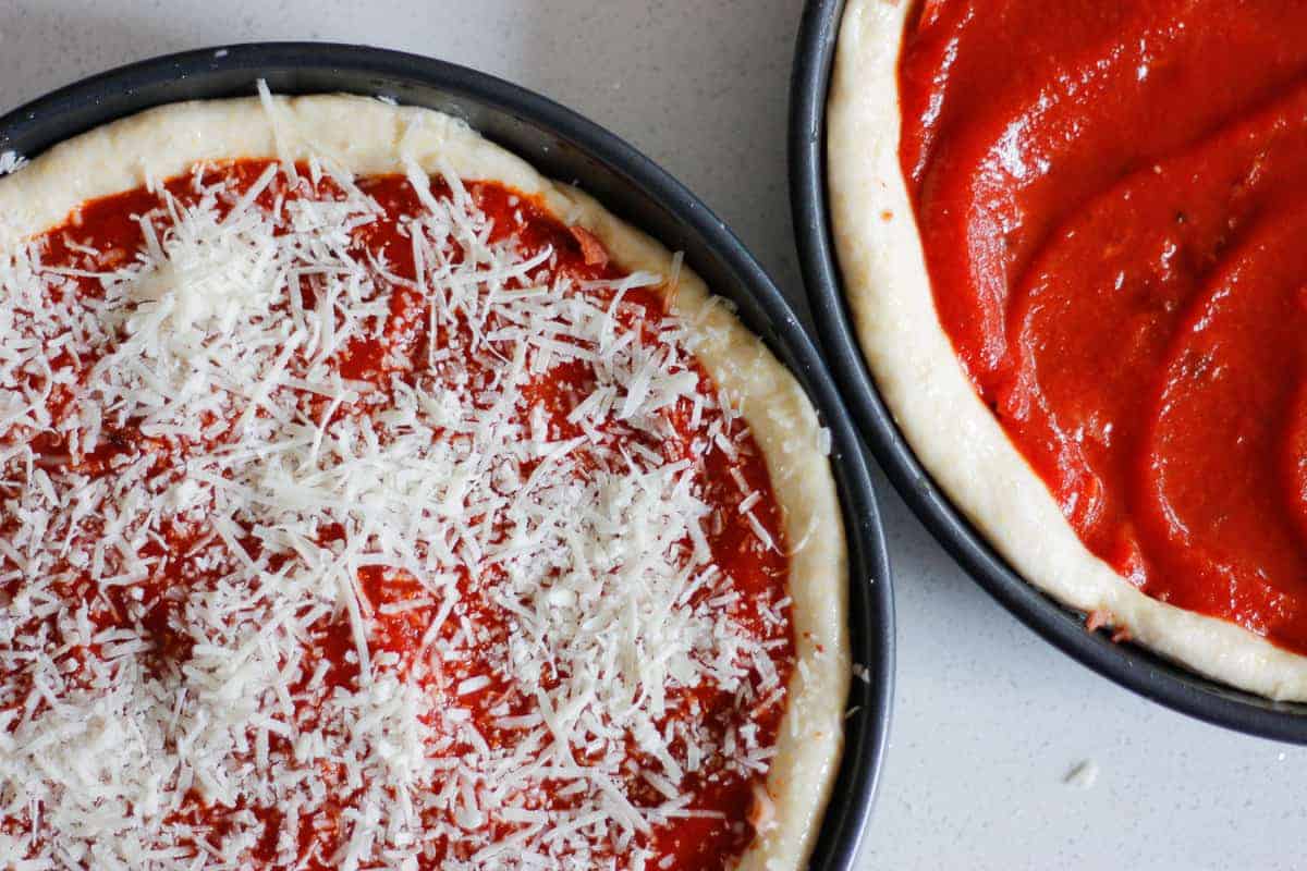 Two Chicago Pizza bases in pans - one with tomato sauce layer, the other with final parmesan sprinkle.