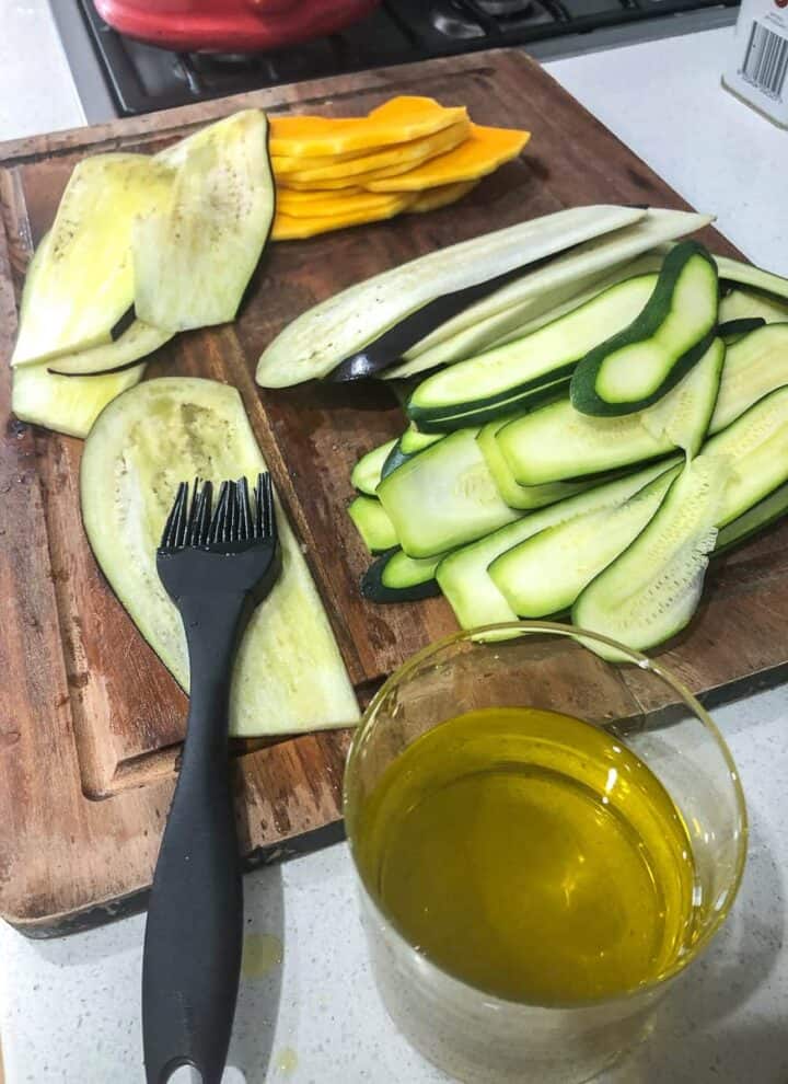 Piles of thinly sliced eggplant, zucchini and pumpkin on a wooden chopping board. A glass of olive oil and a silicone brush ready to coat the slices.