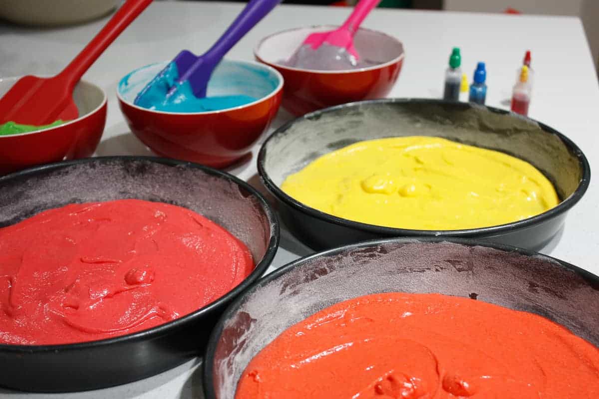 Red, yellow and orange batter in respective tins ready to bake. The remaining three batter colours are in red bowls in the background.