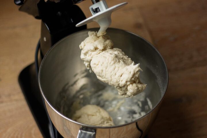 Rough unkneaded dough clinging to the dough hook in a stand mixer bowl.