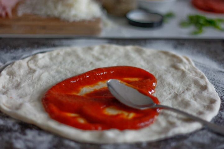 Pizza base being spread with tomato sauce with the back of a metal spoon.