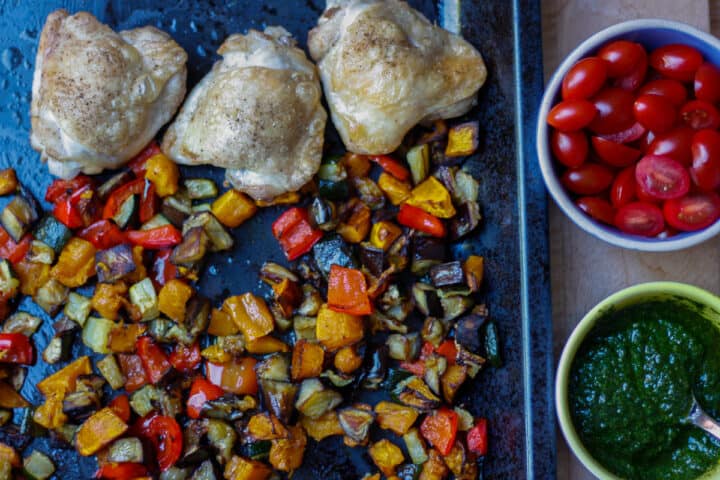 Looking down onto a baking sheet covered with bite-size colourful browned veggies and roasted chicken thighs. Small bowls of basil sauce and chopped cherry tomatoes to the side.