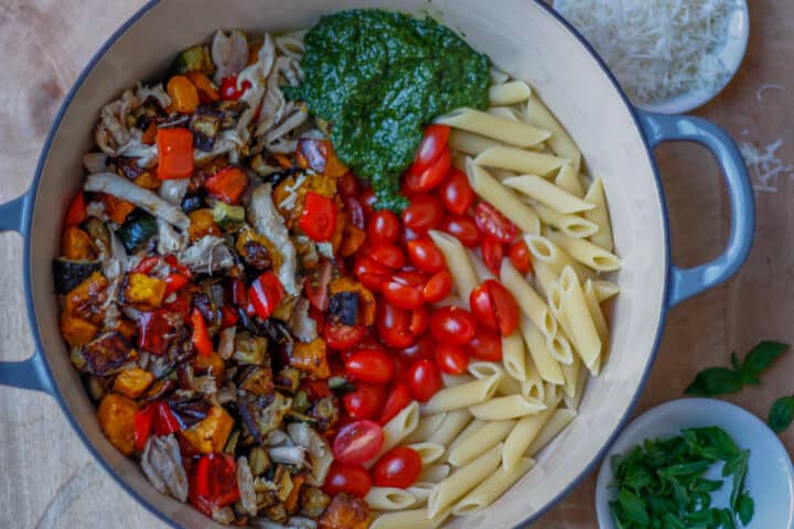 Big pot with separate sections of penne, roasted veg and chicken, cherry tomatoes and basil sauce ready to be combined together.