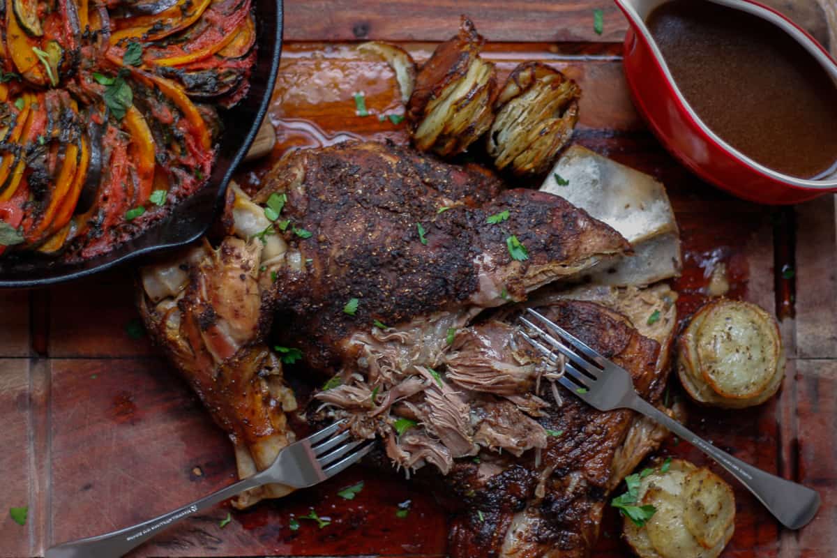 Partially shredded lamb on a wooden board surrounded by sides - ratatouille tian, potato stacks and gravy.