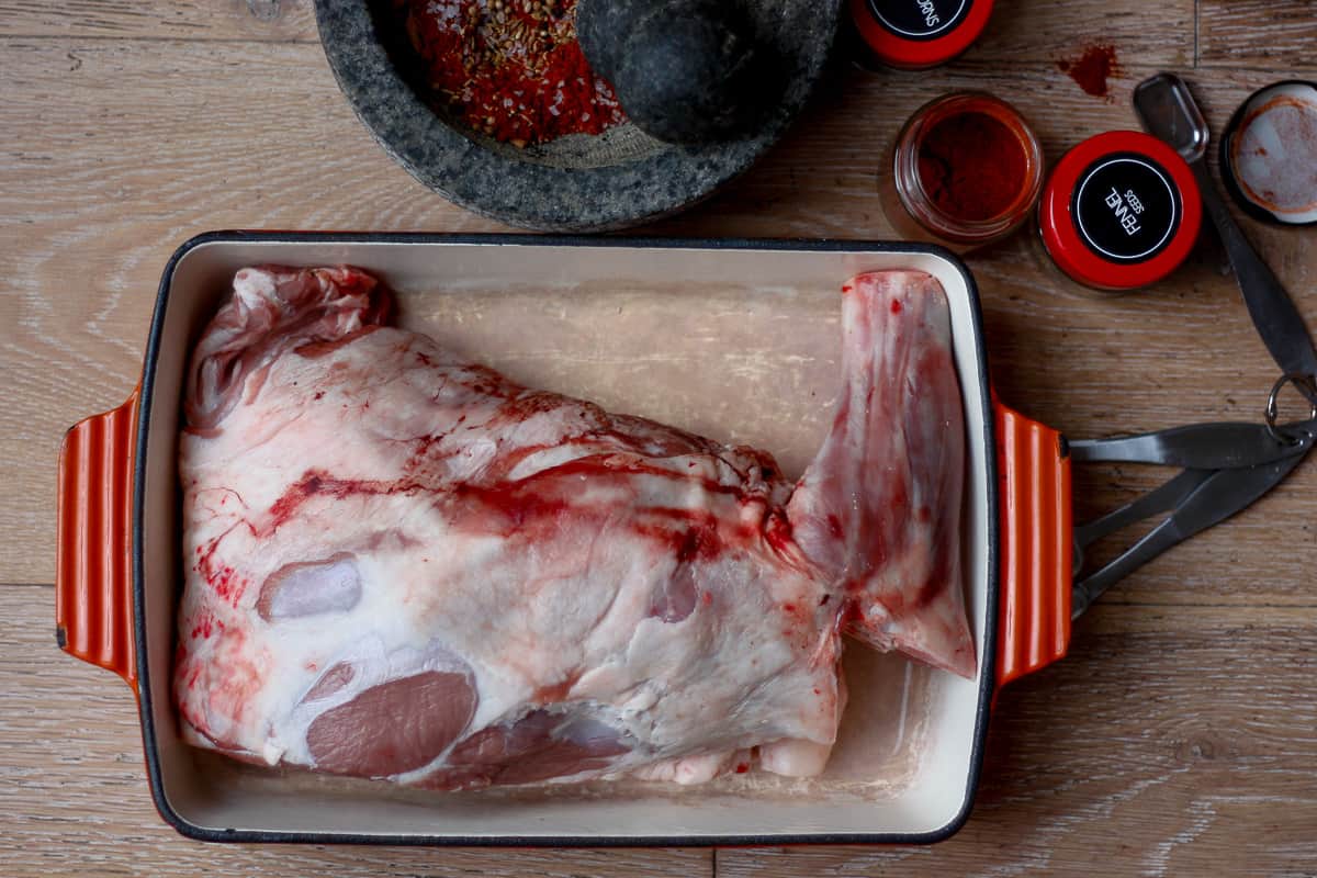 Overhead shot of raw lamb shoulder in a baking dish. A pestle and mortar containing a dark red spice mix is at the upper edge.