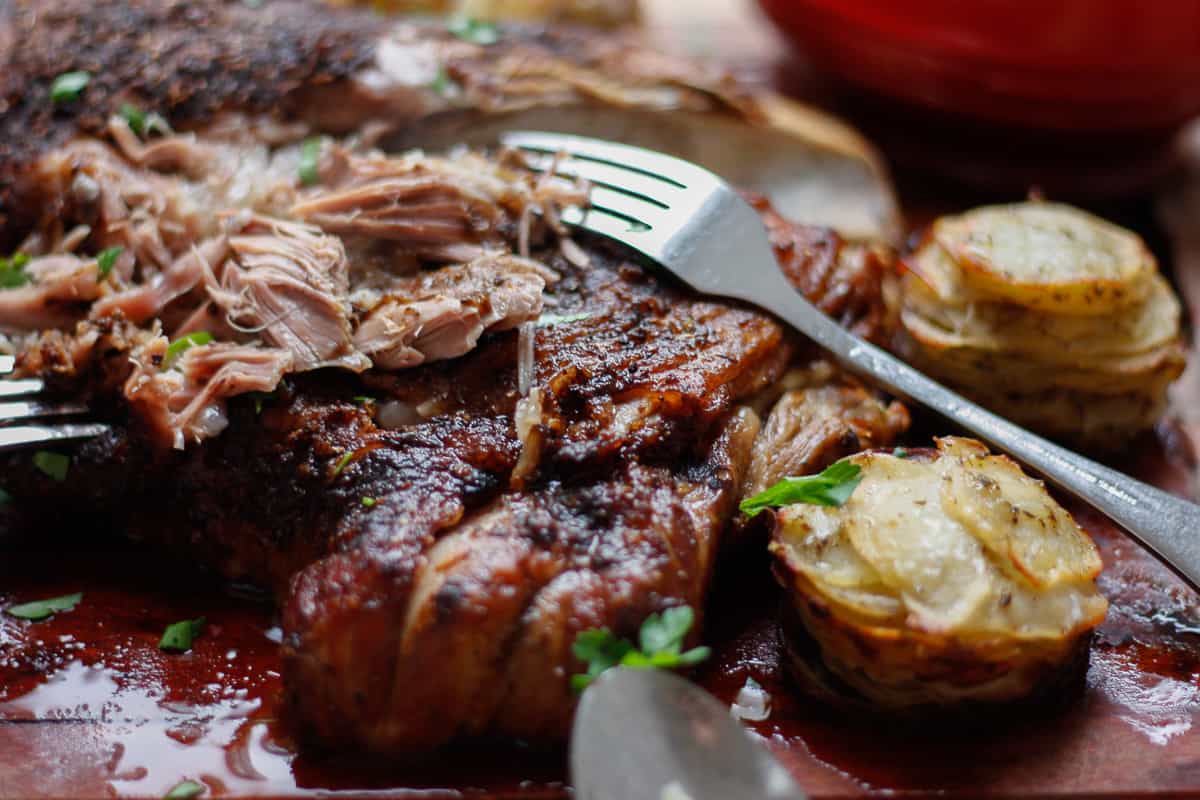 Closeup of lamb partially shredded using two forks. A couple of roasted potato stacks to the side.