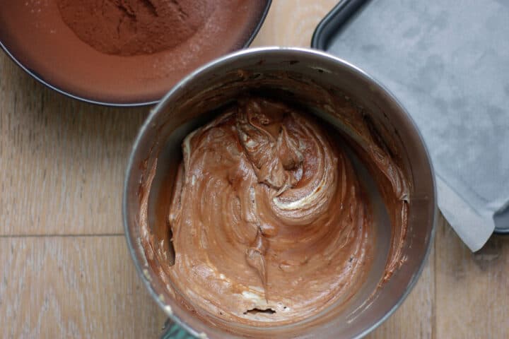 Chocolate coloured wet ingredients combined in a bowl with the dry ingredients in the background.