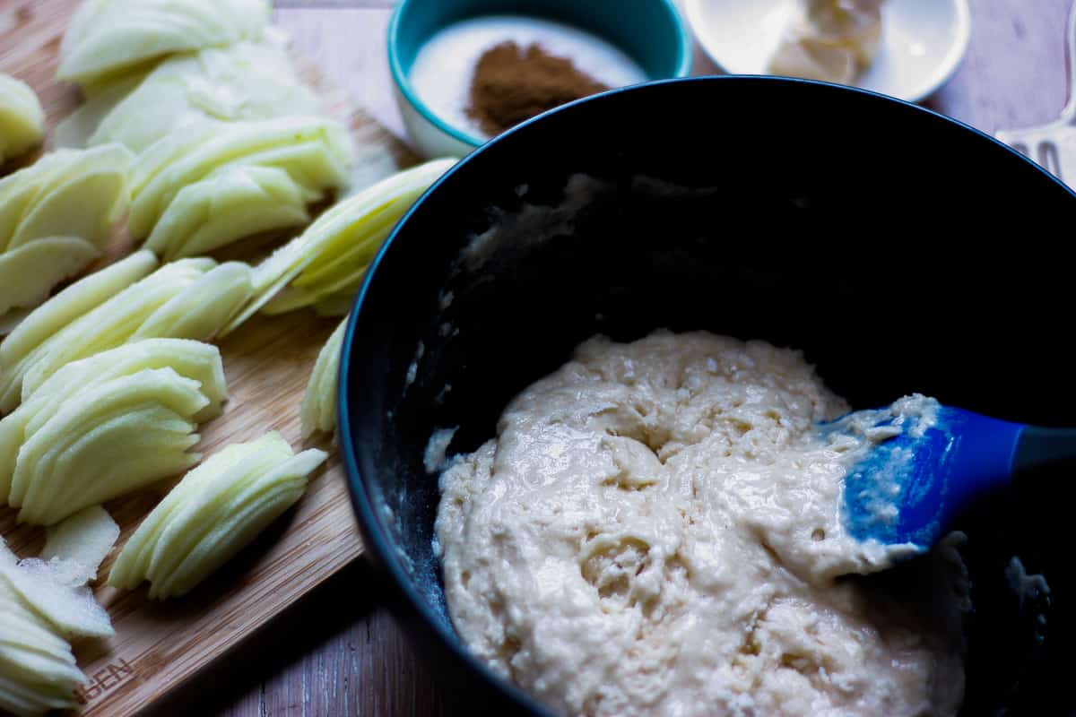 Thick bubbly batter in a black bowl. Groups of thinly sliced apple in a chopping board to the side.