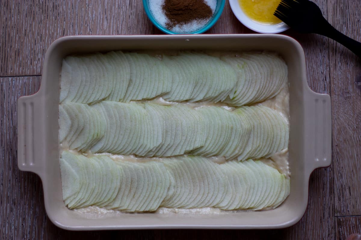 Top view of 3 rows of overlapping thin slices of apple on batter in a baking dish.