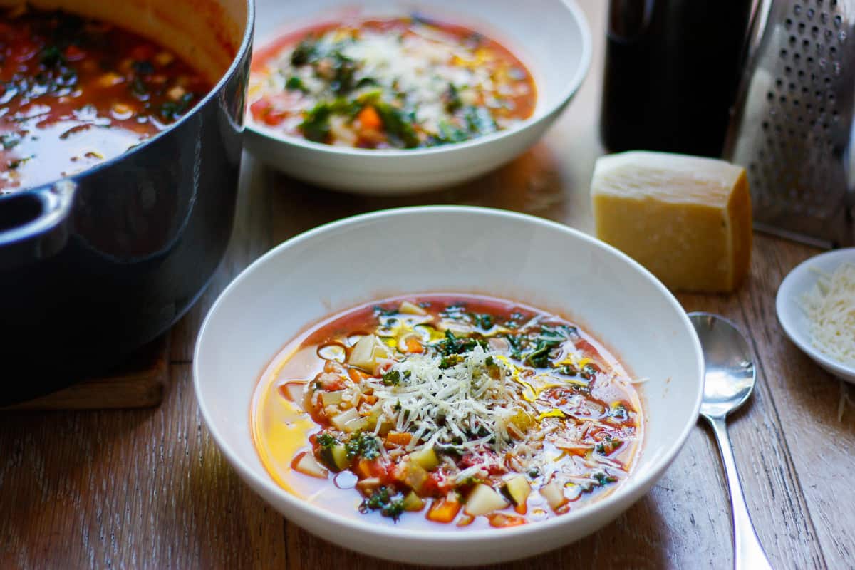 A table set for dinner for two. Minestrone soup in a white bowl topped with grated parmesan in the foreground. The second bowl behind is out of focus.