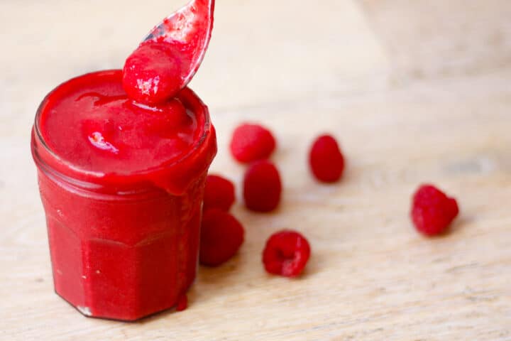 teaspoon dipping in a over-filled jam jar containing finished raspberry sauce. Some sauce dripping down the side.