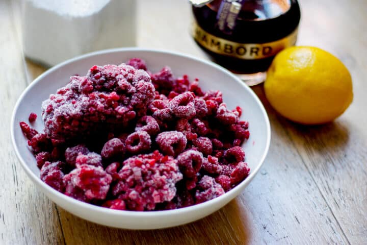 Frozen raspberries in a white bowl with the other ingredients for the sauce in the background - lemon, Chambord and sugar.