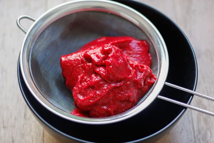 Thick pureed raspberry in a sieve over a black bowl.