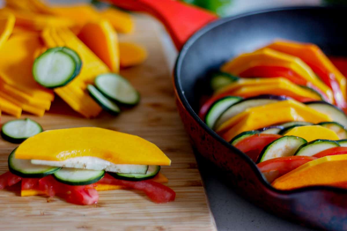 stack of sliced vegetables alongside already layered veg in a pan.