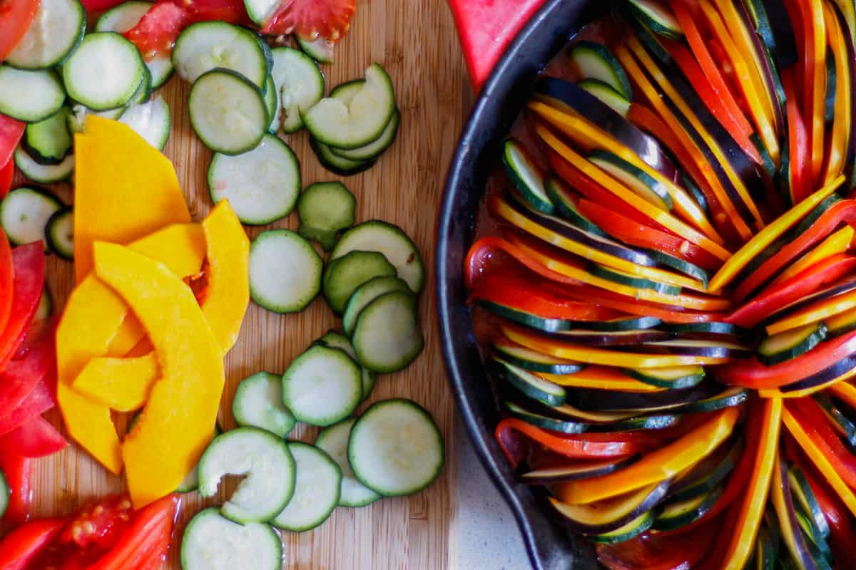 Sliced vegetables on a wooden board alongside a cast iron pan with the veg arranged inside.