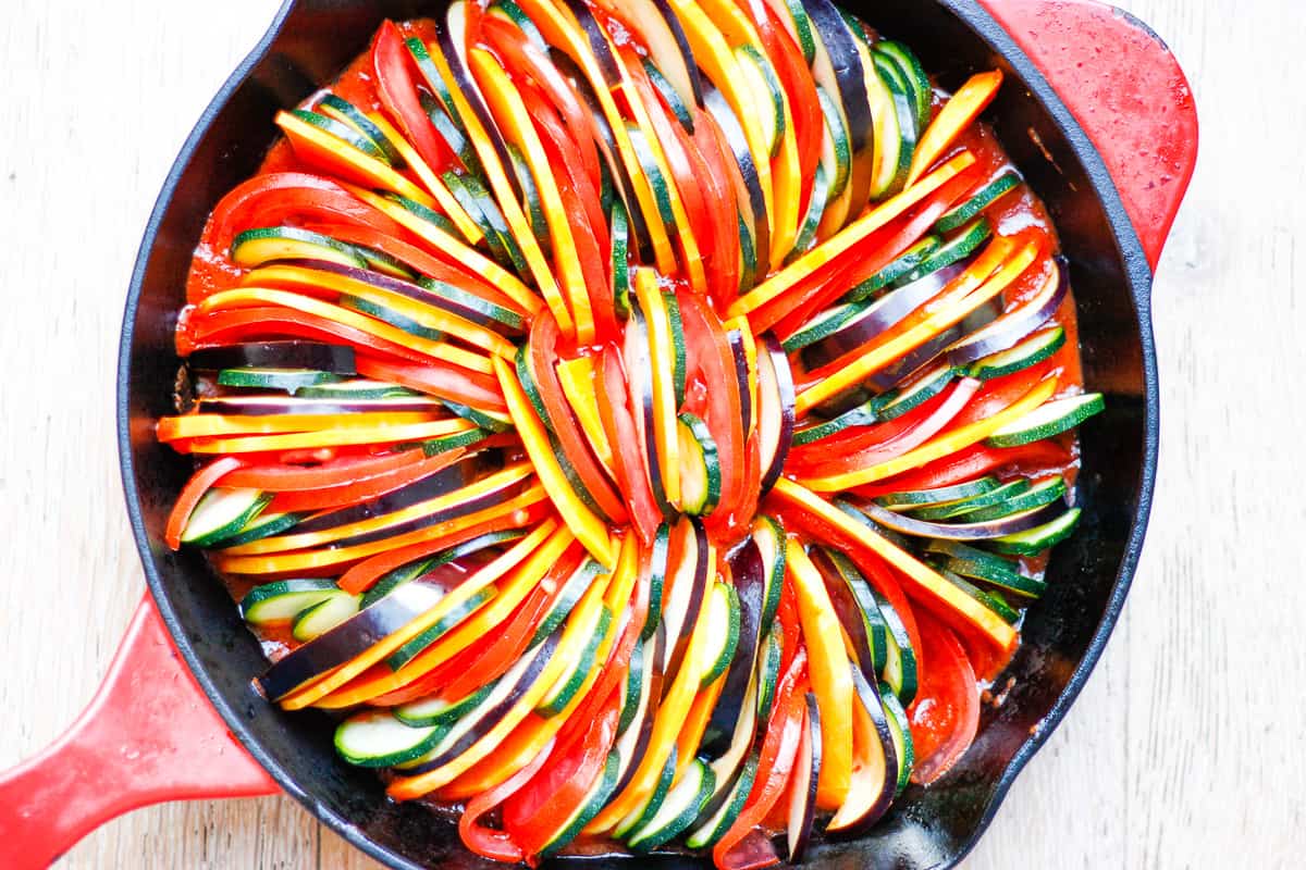 brightly-coloured raw pumpkin, tomato, zucchini and eggplant slices layered in a cast iron pan with a red lid.