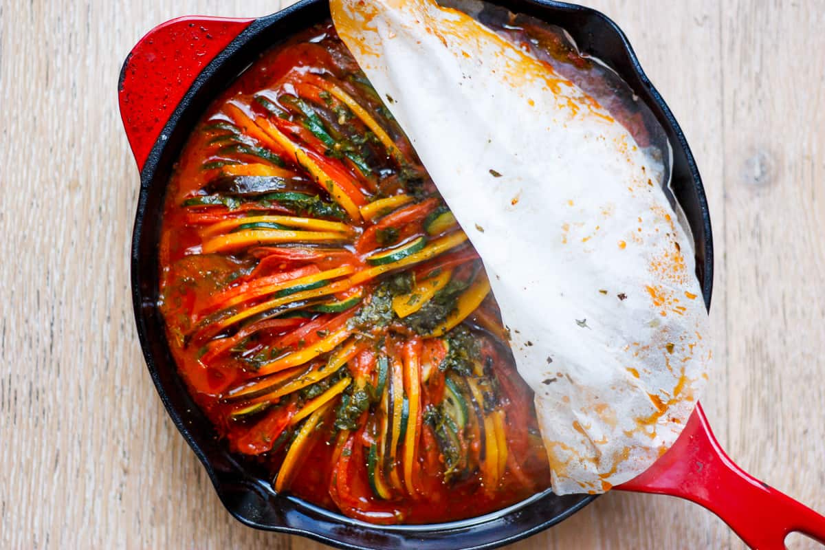 cast iron pan with a red handle. A layer of baking paper partially peeled back to reveal the cooked tian underneath.