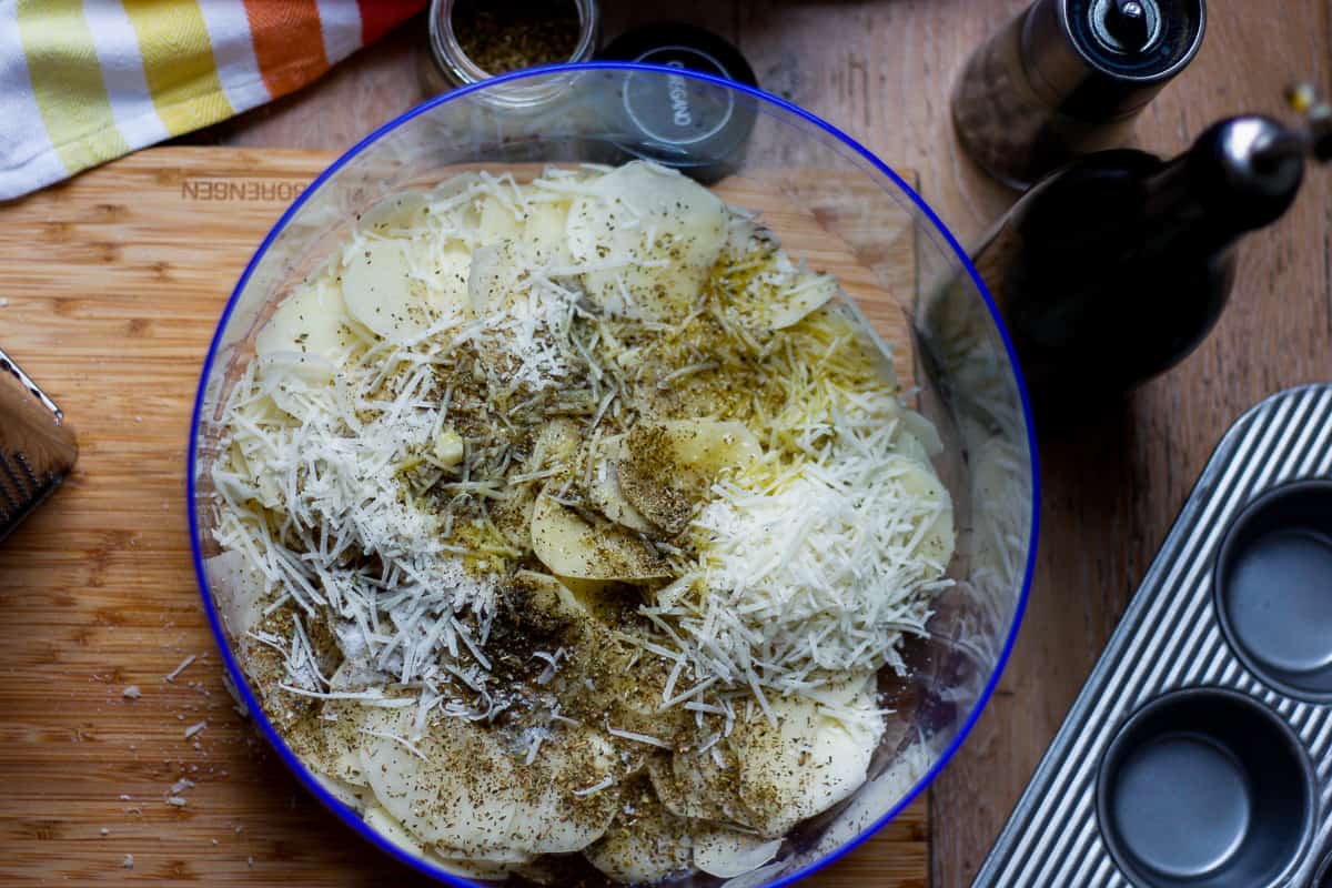 Sliced potato in a large bowl covered in seasoning and grated cheese.