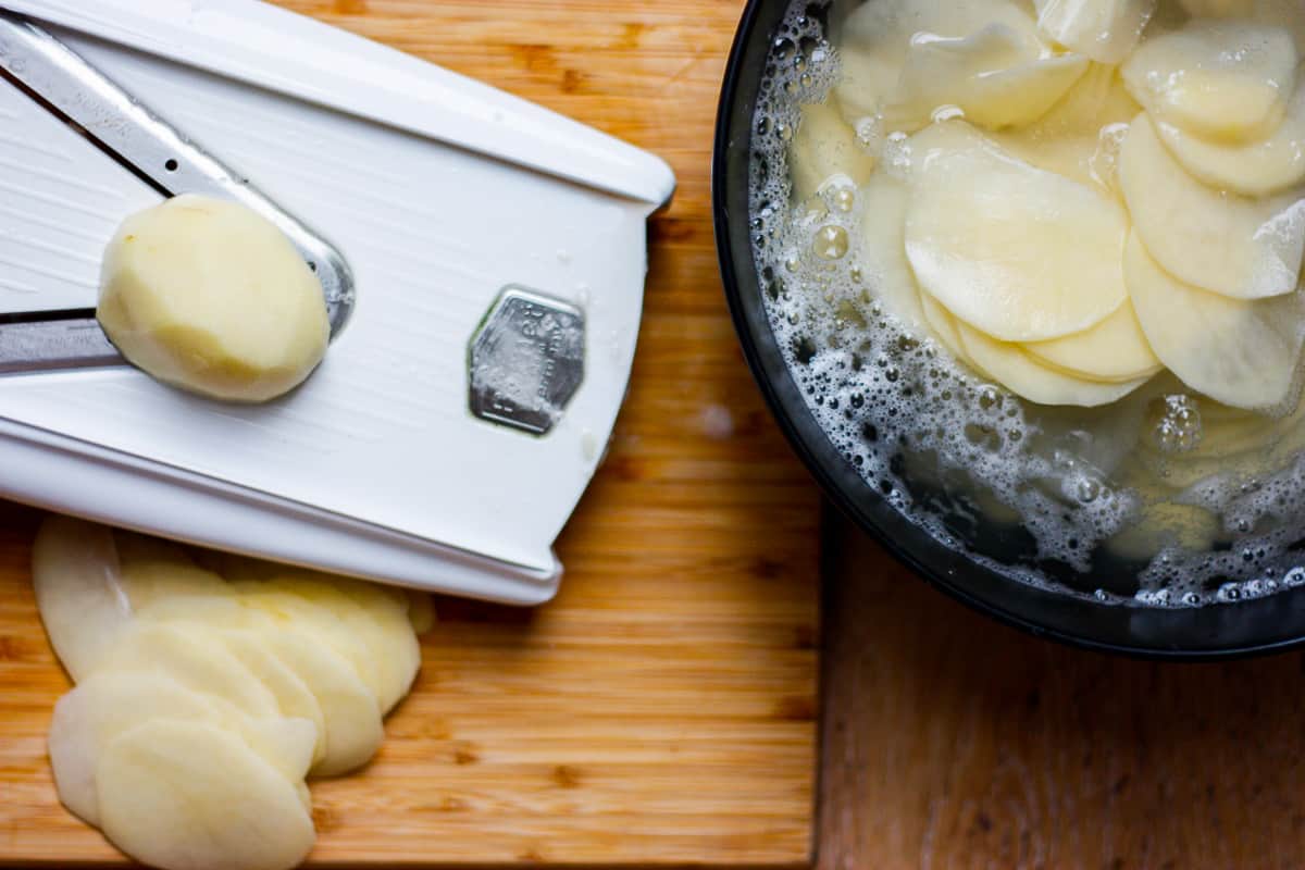 A partially-sliced potato resting on a mandolin. Already sliced potato in starchy water in a bowl to the side.