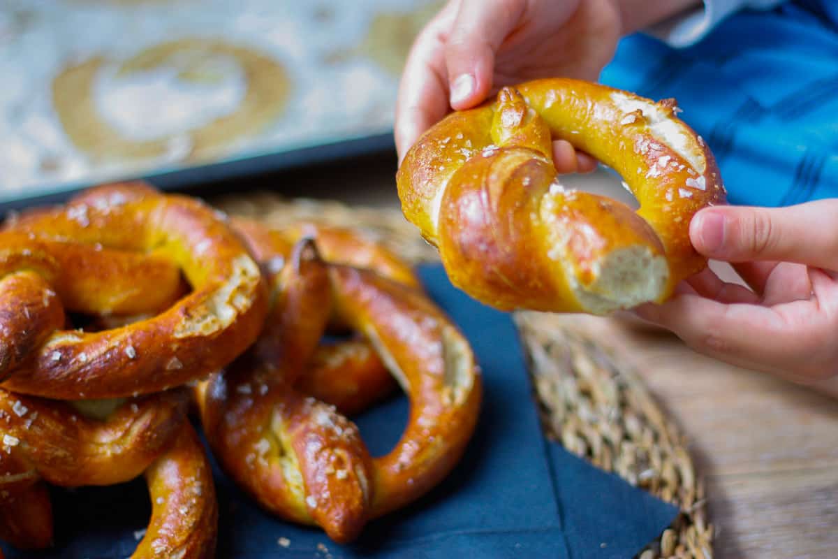 A pretzel held between a young child's hands. A pile of pretzels on blue napkins set on a wicker place mat.