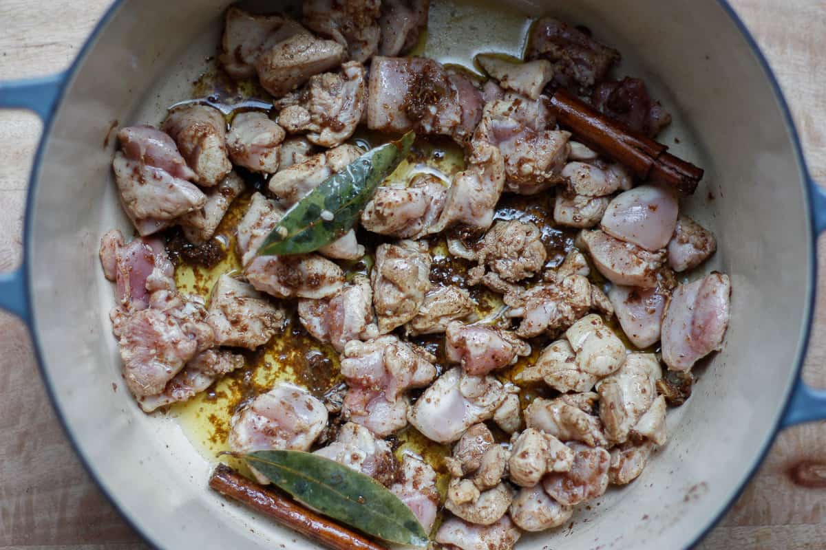 Bite-size pieces of chicken coloured slightly in a pot melted spiced ghee. Bay leaves and cinnamon sticks resting on top.