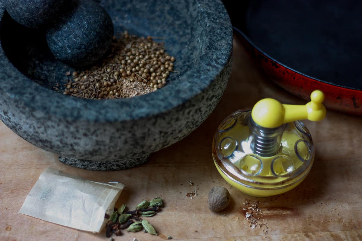 Some of the whole spices used in Chicken Pilau - cloves and split cardamon pods tumbling out of a spice bag, a nutmeg grinder with a yellow handle and a mortar containing coriander and cumin seeds.