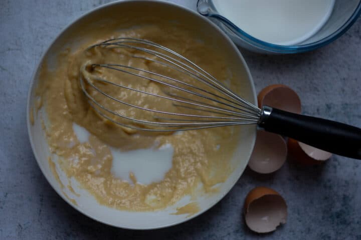 Milk being gradually added to the batter with a large handheld whisk.