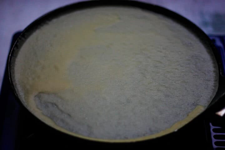 Thin layer of batter cooking in crepe pan with bubbles of condensation on the surface and edges starting to curl.