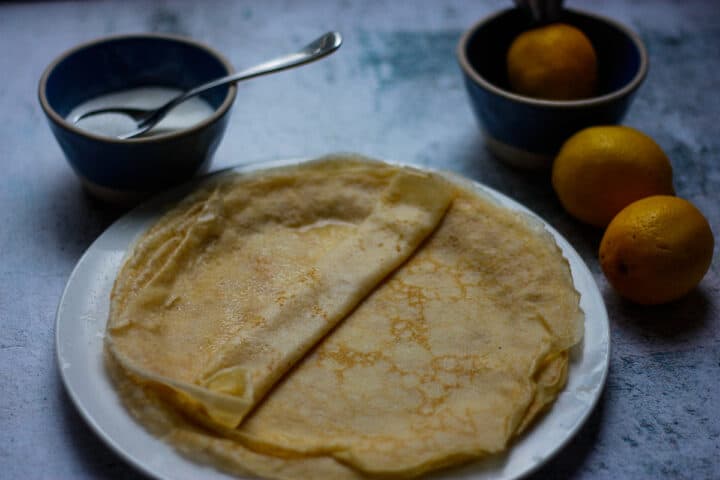 Stack of crepes on a plate. Lemons and sugar in the background.