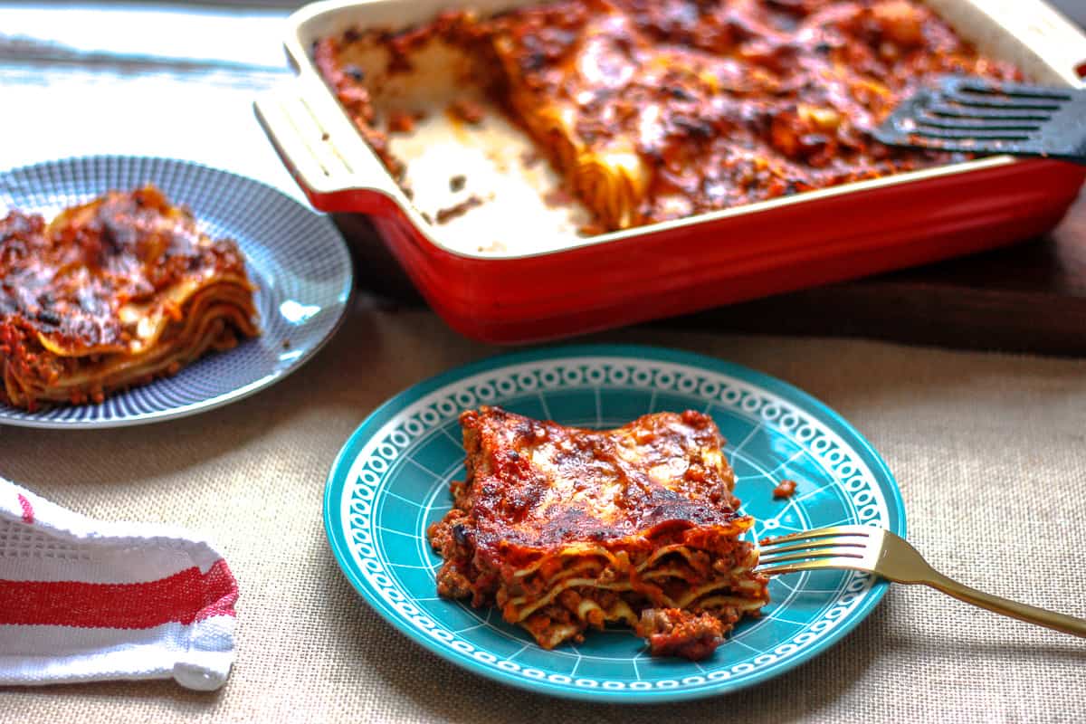 Generous portion of lasagna on a small blue plate. There's another serving in the background and the whole lasagna with two pieces missing.