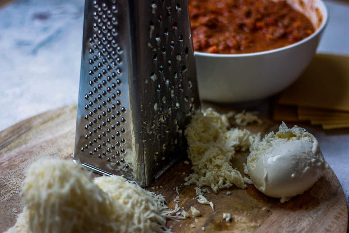 Piles of grated parmesan and mozzarella around a box grater. There's a big bowl of bolognese sauce in the background.