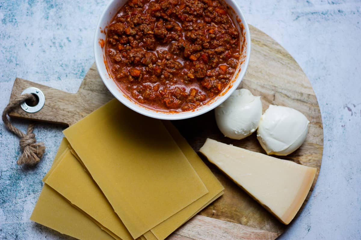 Ingredients to make lasagne - bolognese sauce, mozzarella balls, triangular wedge of parmesan and lasagne sheets. All set on a round wooden chopping board.