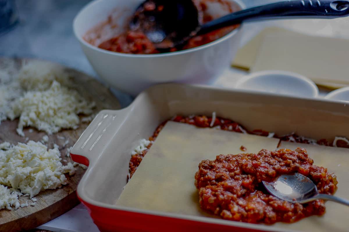 Demonstrating the layering process. A portion of bolognese sauce being spread on a layer of lasagne sheets in a large baking dish.