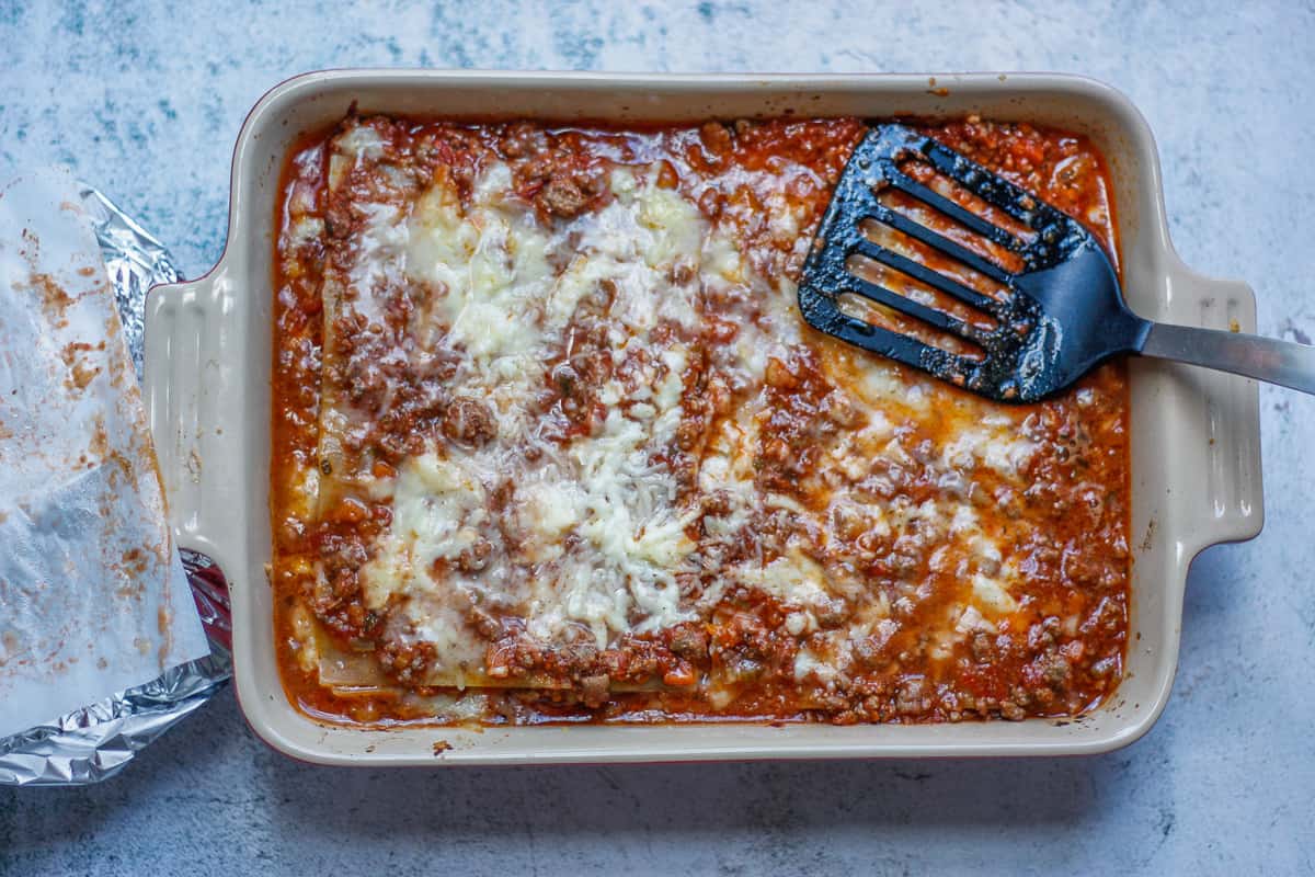 Half-baked lasagne. The baking paper and foil layers are off to the side of the baking dish. A fish slice is being used to push the lasagne sheets down.