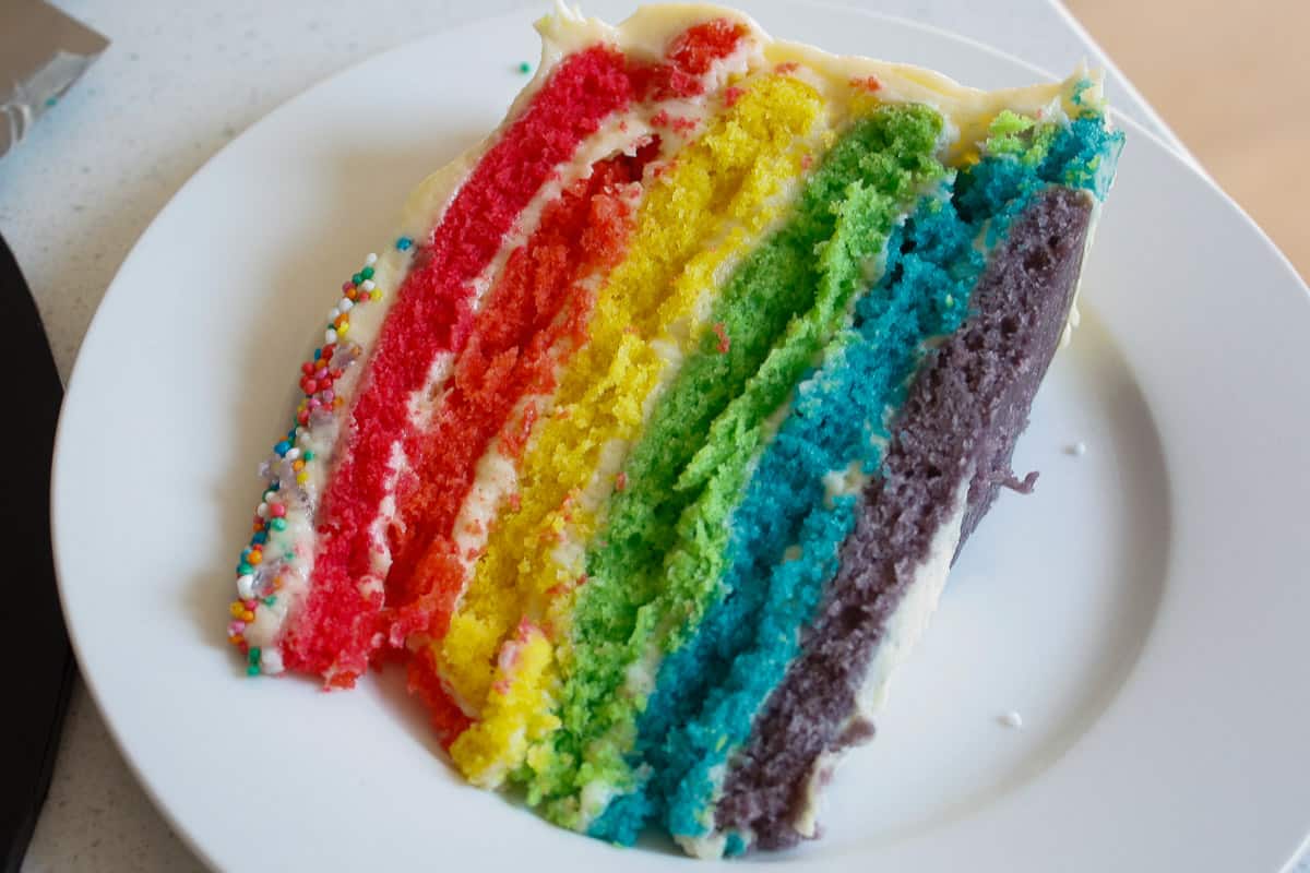 Slice of rainbow cake lying on its side on a white side plate. You can see all the layers with frosting between them.